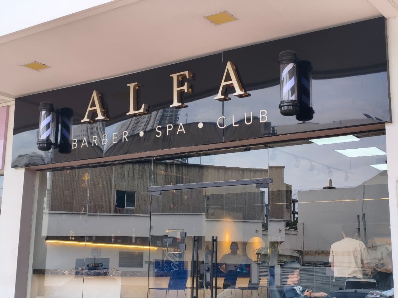 Fachada de ALFA Barber Spa Club: el letrero negro con letras doradas y los dos postes de barbería a los lados